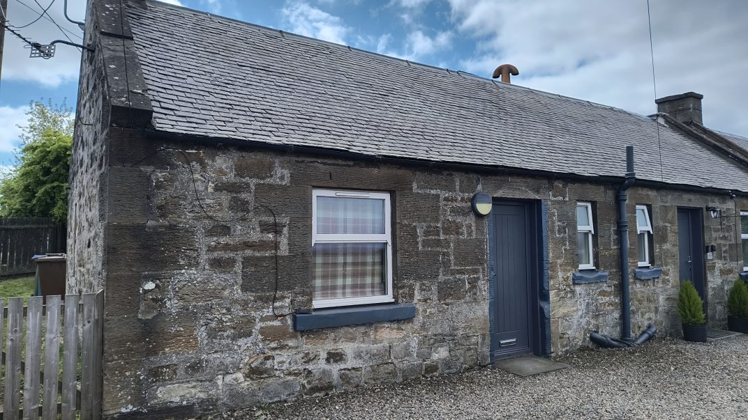 Stone cottage with a slate roof, exposed stone walls, blue doors, and a gravel yard; a round external light sits between the window and door, with black downpipes along the wall.