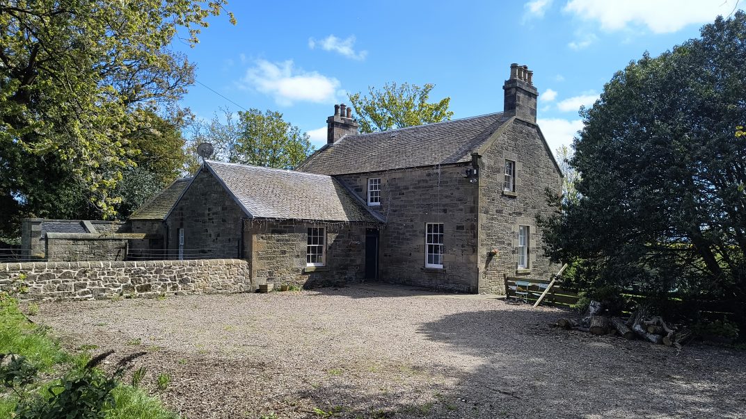 Stone stone-faced cottage with a slate roof and two chimneys, set in a gravel yard surrounded by trees and blue sky above.