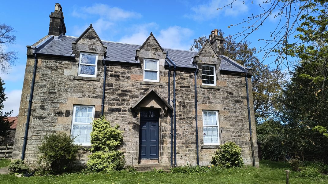 Front view of a gray stone house with a dark blue door, slate roof, and dormer windows under a blue sky, surrounded by greenery.