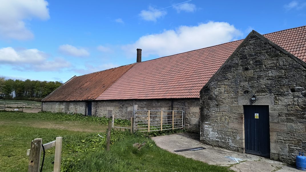 Stone barn with a red tiled roof and chimney under a blue sky, in a rural field with a wooden fence nearby.