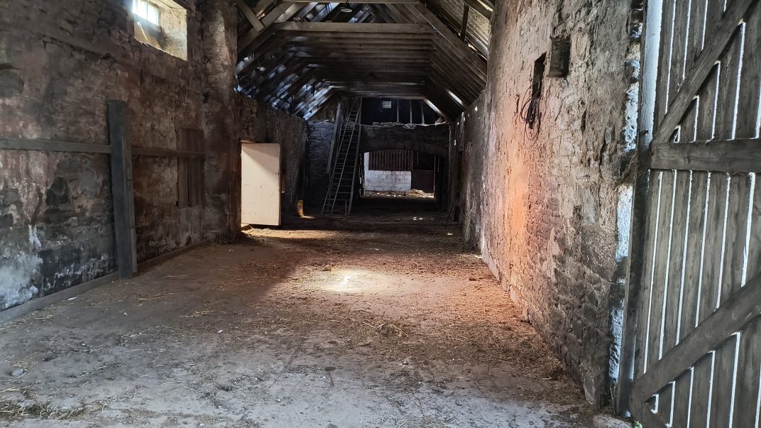 Dim barn interior with stone walls, wooden beams, and a ladder to the loft at the far end.